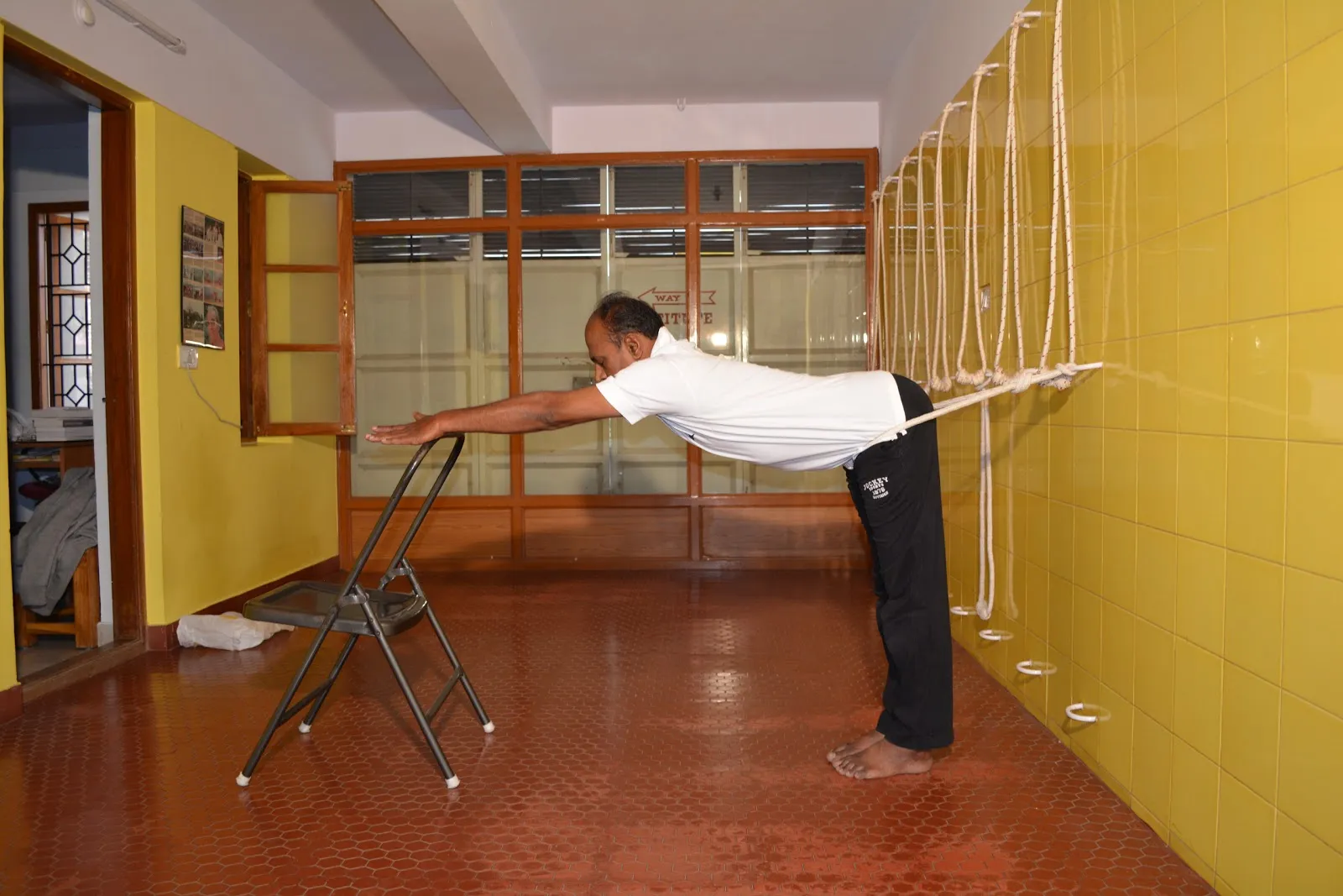 Narayan Sir in a forward extension in the hall, hands on a chair and a rope around his hips, with ropes hanging on the yellow tiled wall